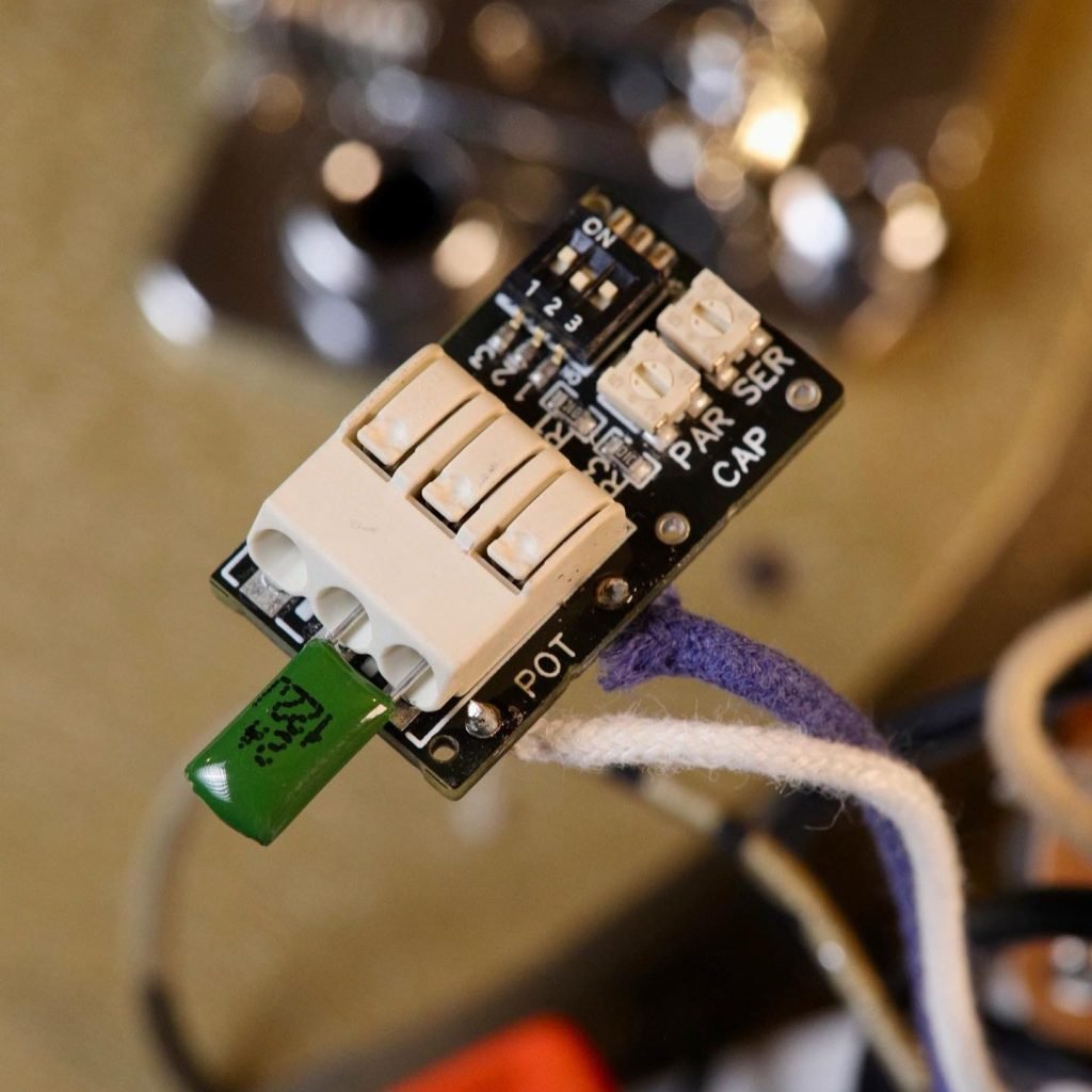 Close-up of the Mars-Tronic Treblemaker with a visible capacitor in push-fit terminals and blue-and-white cloth wires, against a blurred backdrop of guitar electronics.